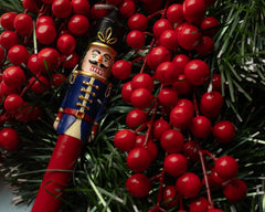 Sir Elliott nutcracker candle with a navy blue jacket and red stem nestled among red berries and evergreen branches.
