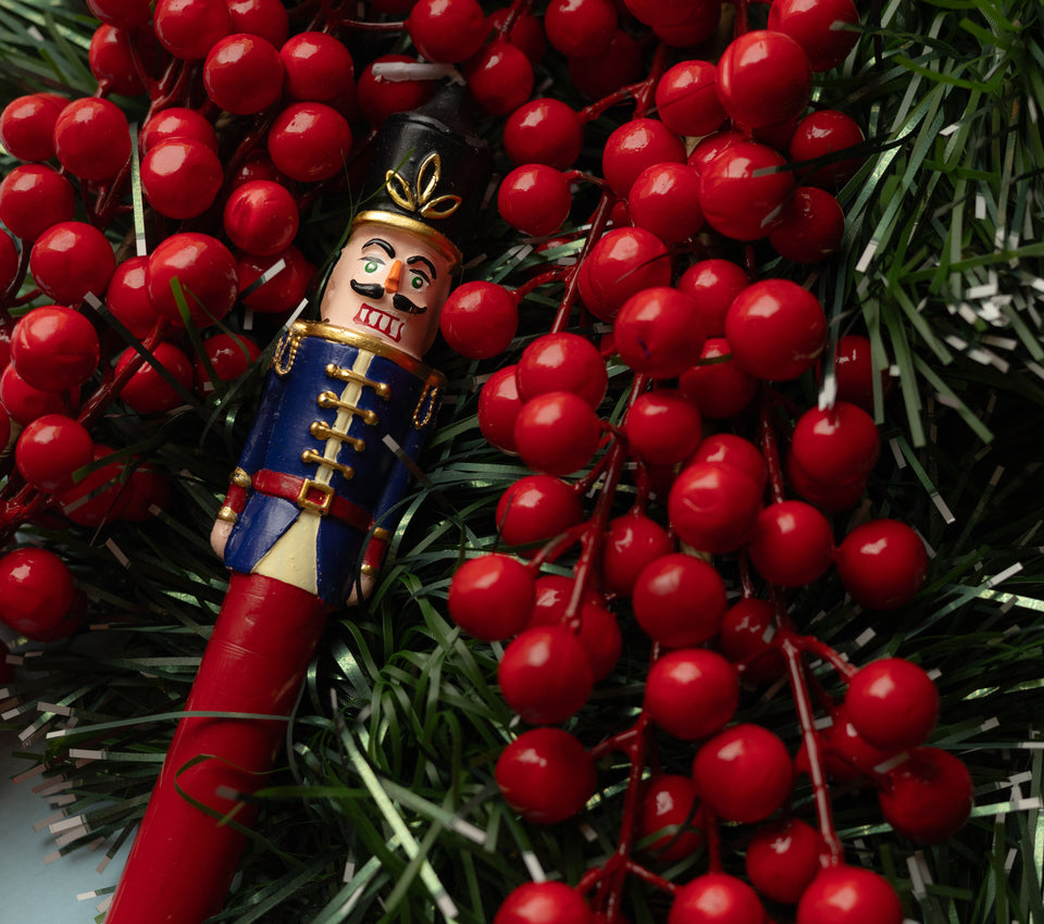 Sir Elliott nutcracker candle with a navy blue jacket and red stem nestled among red berries and evergreen branches.
